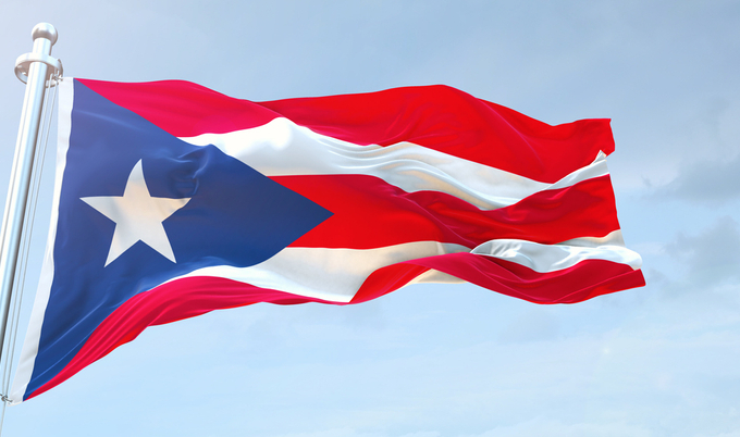 A closeup of the Puerto Rican flag on a flagpole, waving in the wind with a light blue sky in the background.