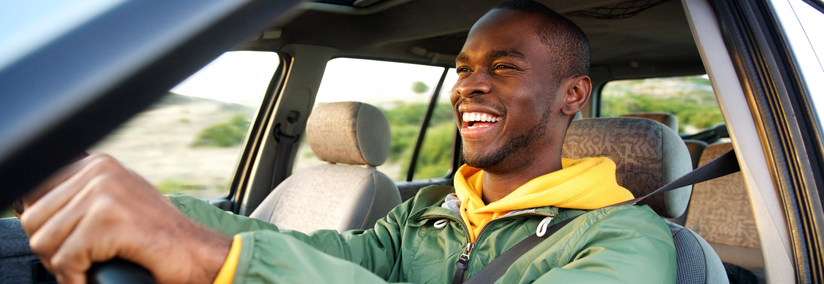 A smiling young man is sitting in the driver's seat of a car with his hands on the steering wheel.